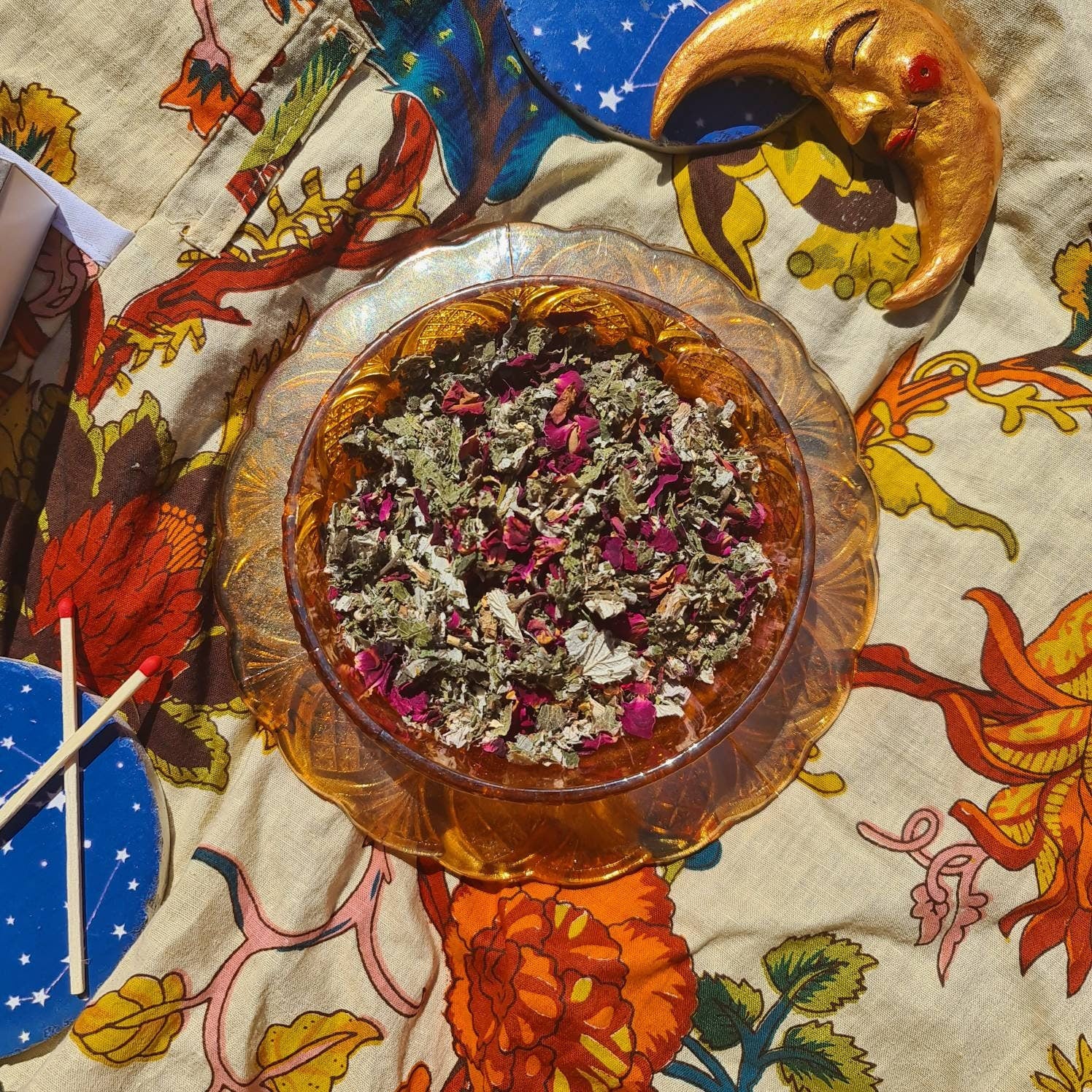 An enchanting image of The Dreamy Blend in a carnival glass bowl showcasing a blend of green raspberry leaf, mugwort, rose petals, and lavender. A celestial moon decoration with twinkling stars adorns the background, adding a touch of magic.