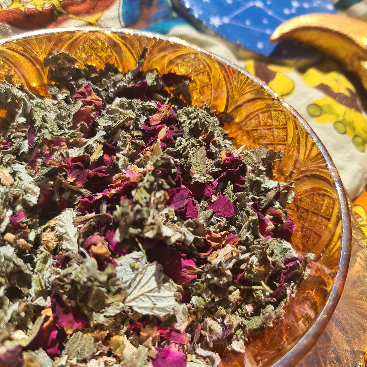 An enchanting close up image of The Dreamy Blend in a carnival glass bowl showcasing a blend of green raspberry leaf, mugwort, rose petals, and lavender. A celestial moon decoration with twinkling stars adorns the background, adding a touch of magic.